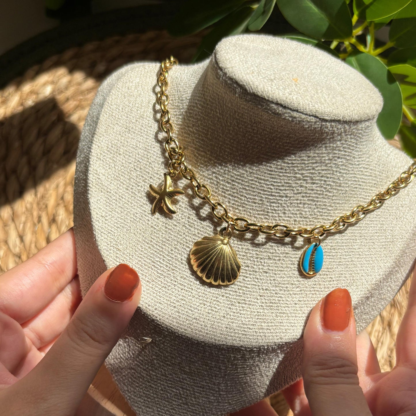Gold necklace with charms on a white bust, held by hands with orange nail polish.