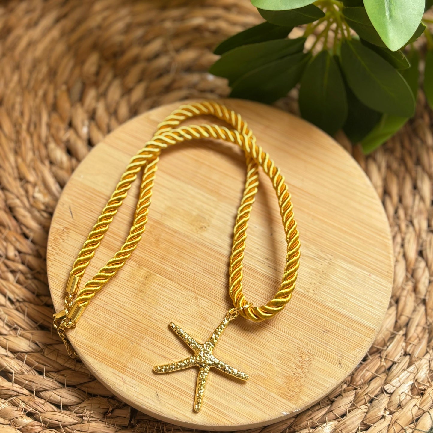 Gold necklace with a starfish pendant on a wooden coaster, surrounded by green leaves.