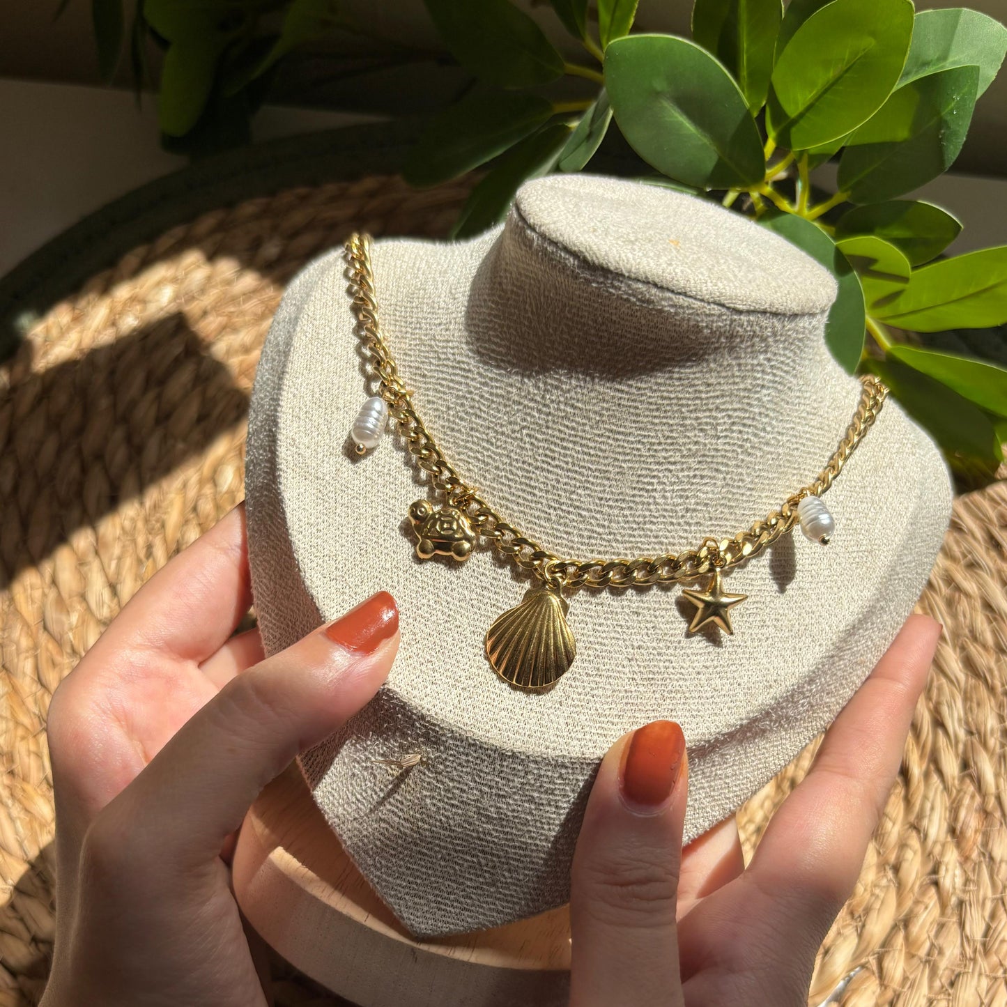Gold necklace on a bust held by hands with orange nail polish, against a natural background.